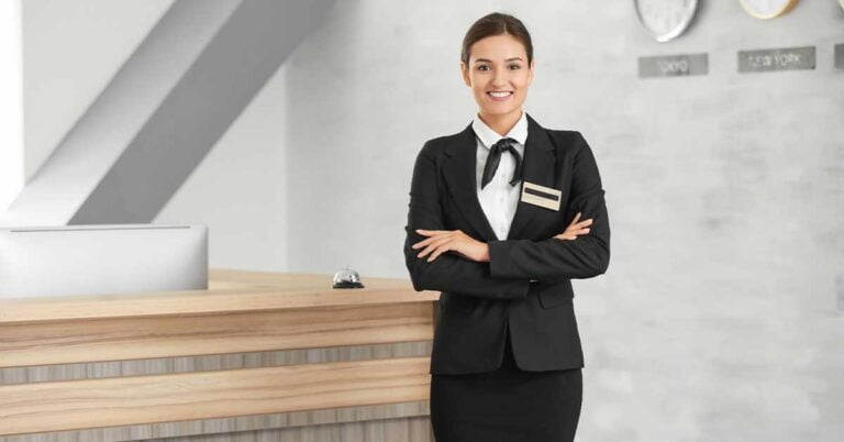 A female hotel receptionist stands smiling at the reception desk.