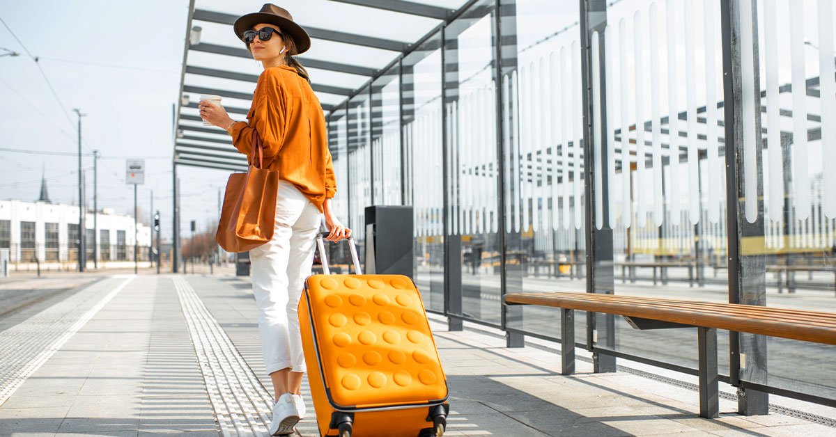 a woman wearing white pants and orange shirt with a brown hat and sunglasses dragging a suitcase