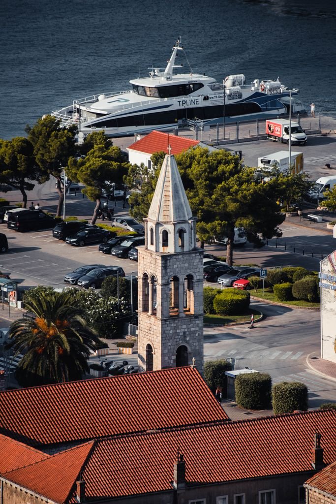 Photo by Ajdin Coric A picturesque view of Herceg Novi's harbor featuring a prominent church tower and boats docked by the seashore.